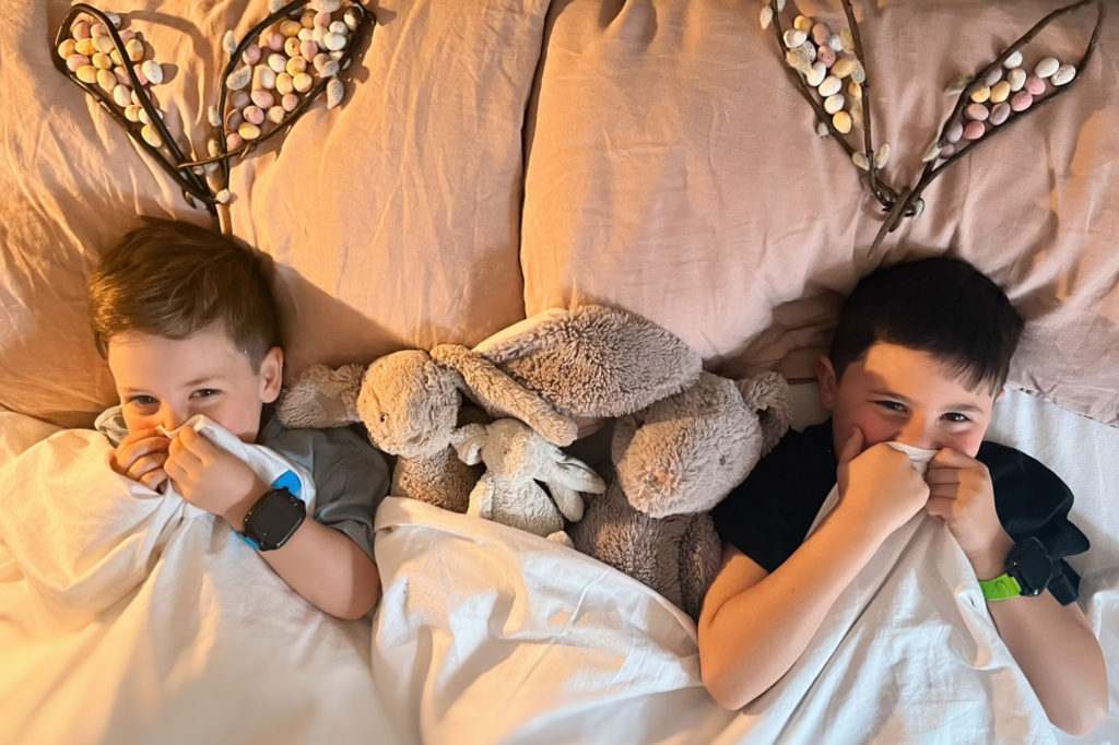 en familie på fire med to små brødre der hver har et xplora mobilur der er lagt under dynen. Fra venstre er der en dreng med et blå ur, og til højre en dreng med med et limegrønt ur.